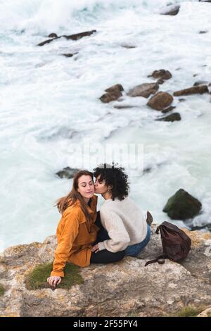 Giovane donna che bacia la guancia dell'amico mentre si siede sulla scogliera via mare Foto Stock