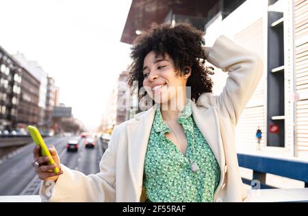 Giovane donna con i capelli a mano che ascolta la musica tramite Bluetooth auricolari mentre si utilizza uno smartphone in città Foto Stock