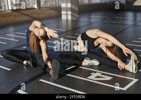 Due attraenti atleti sportivi, donne sportive in abito da yoga, seduta sul pavimento della palestra, stretching prima dell'allenamento mattutino, riscaldamento per iniziare produttivo Foto Stock