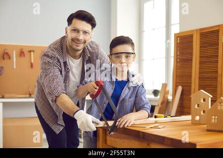 Padre e figlio indossano occhiali di protezione durante il taglio della tavola di legno presso l'officina del falegname Foto Stock