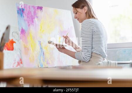 Giovane donna che tiene la tavolozza di colori mentre dipingendo tela astratta a. casa studio Foto Stock