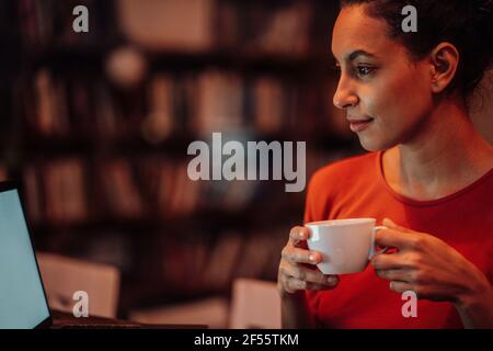 Donna sorridente che beve caffè mentre usa il computer portatile seduto al bar Foto Stock