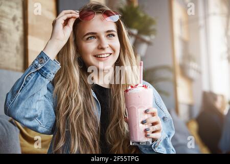 Ragazza che vede faccia familiare sulla strada mentre si siede in un locale cafè all'aperto, prendendo degli occhiali da sole per dare un'occhiata, tenendo un cocktail, invitando un amico a. Foto Stock