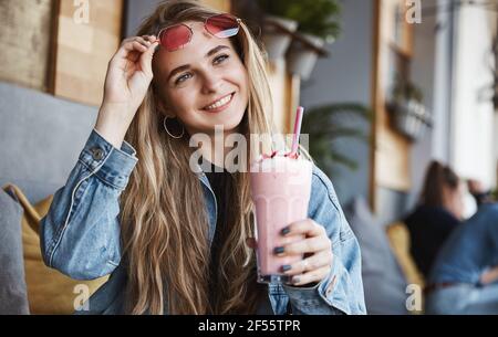 Happy ragazza take-off occhiali da sole e frullato bere nella terrazza caffetteria, guardando fuori dalla strada. Donna bionda con cocktail di fragole al sole Foto Stock