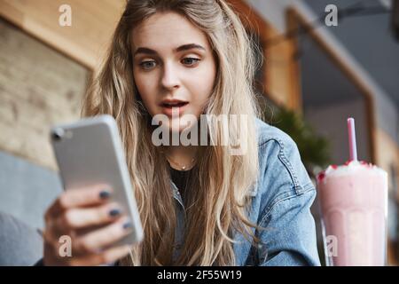 Primo piano di una ragazza entusiasta che guarda lo schermo dello smartphone in un caffè all'aperto, bevendo frullato e leggendo un messaggio telefonico Foto Stock