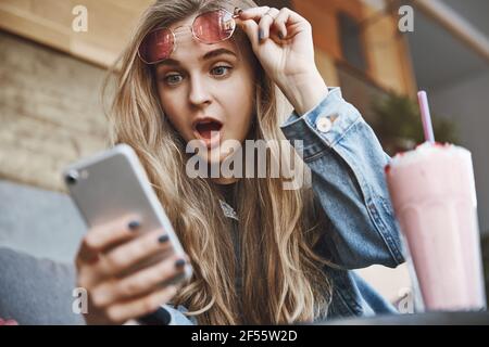 Lol, nessun modo. Ritratto di una studentessa europea in denim emozionale e attraente, che tesse gli occhiali da sole rosa mentre si sta guardando lo schermo dello smartphone Foto Stock