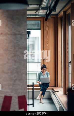 Donna imprenditore che lavora su un computer portatile mentre si siede sopra la finestra alcova posto in ufficio Foto Stock