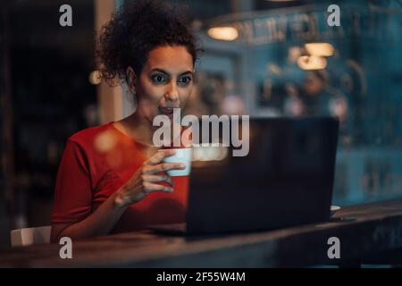 Giovane donna che tiene la tazza di caffè mentre fissando al computer portatile con bocca aperta nella caffetteria Foto Stock