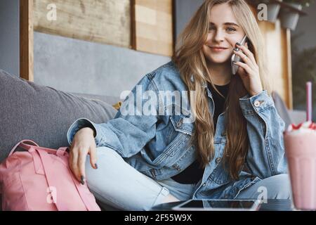 Ragazza che chiama il taxi, seduto con gli amici e che vuole guidare a casa, seduto sul divano con i piedi incrociati, tenendo lo smartphone vicino all'orecchio, sorridente spensierato Foto Stock