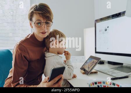 Mamma che lavora con lo smartphone mentre il figlio guarda i video tablet digitale Foto Stock