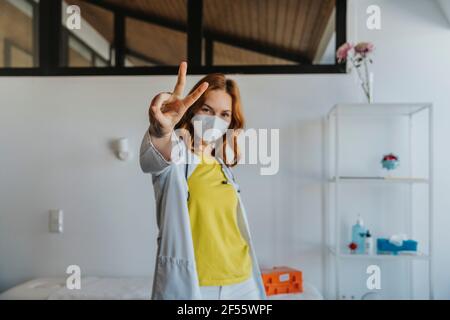 Medico indossando maschera protettiva viso stretching mano mentre gesturing pace cartello in piedi presso la clinica Foto Stock