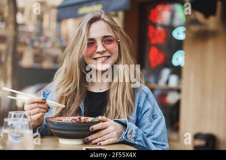 Felice imprenditore femminile in occhiali da sole alla moda e giacca in denim, guardando da parte con sorriso soddisfatto, essere impressionato con il gusto sorprendente del cibo asiatico Foto Stock