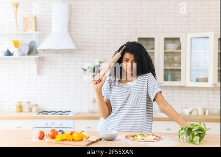 Triste incerto chef novizio donna latina cercando di fare una pizza italiana originale a casa, tenendo un tondino vicino alla testa guardando la macchina fotografica, non sicuro del gusto del cibo, è noioso per lei Foto Stock