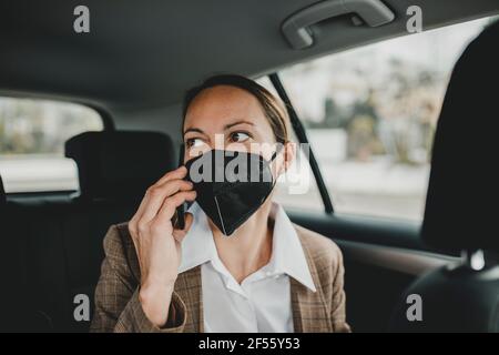Donna d'affari che indossa una maschera protettiva che parla su smartphone mentre viaggio in taxi Foto Stock