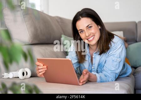 Bella donna sorridente mentre tiene il tablet digitale sul divano casa Foto Stock