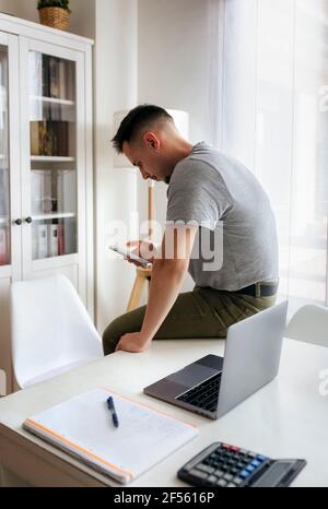 Imprenditore maschile che utilizza lo smartphone mentre si siede sulla scrivania da un computer portatile in ufficio a casa Foto Stock