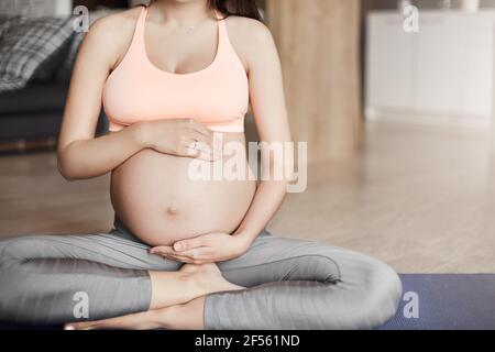 Ripresa di una giovane donna in gravidanza sana in reggiseno sportivo e leggings, seduta in posa yoga sul roll pad, toccando il ventre come se abbracciasse il futuro bambino Foto Stock