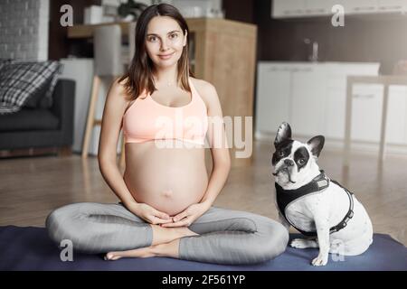 Scatto orizzontale di buona-guardare donna incinta felice, in attesa di diventare mamma, seduta con cane amorevole sul roll pad in posa yoga, toccando la pancia e. Foto Stock