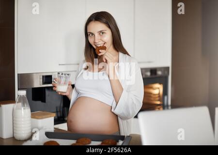 Le donne incinte non condividono il cibo. Moglie europea affascinante e premurosa con il ventre carino, in piedi vicino al tavolo da cucina con biscotti su gocciolatore-padella, mordere Foto Stock