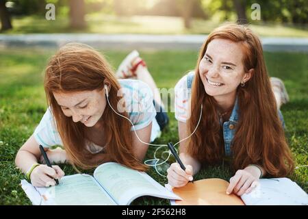 Smettete di ingannare e di fare i compiti. Due rilassate e spensierate sorelle rosse con frettole, adagiate sull'erba nel parco, scrivono note per l'università Foto Stock