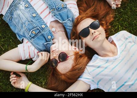 Allegra ragazza rossa sdraiata sull'erba nel parco con il migliore amico, indossando occhiali da sole per nascondersi dal sole, sorridendo con espressione rilassata e spensierata Foto Stock