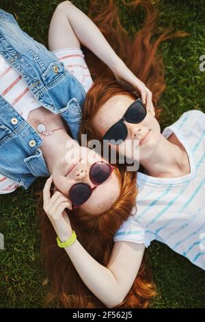 I gemelli sono i migliori. Scatto verticale di due belle sorelle con capelli allo zenzero e fracche, sdraiati sull'erba e sorridenti con espressione rilassata, toccanti Foto Stock
