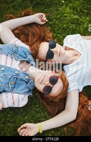 Scatto verticale di belle ragazze rilassate e felici con i capelli di zenzero naturali e carini che giacciono sull'erba, che si toccano l'un l'altro teste, indossarsi fresco Foto Stock