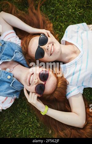 Sorelle gemelle zenzero che indossano sfumature adagiato sull'erba in una soleggiata giornata autunnale godendosi il cielo. Crescere insieme nella follia della testa rossa. Foto Stock