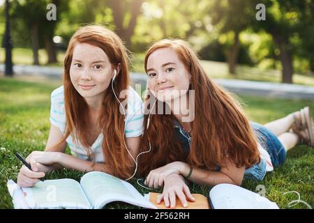 Le ragazze fanno tutto insieme. Affascinanti donne con testa rossa naturale in abiti estivi distesi sull'erba durante i fine settimana, condividendo gli auricolari per ascoltare le canzoni Foto Stock