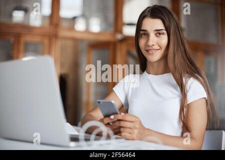 Ritratto di giovane donna che usa il laptop e il telefono cellulare nella biblioteca universitaria preparandosi per i suoi esami di laurea. Giovane professionista che la inizia Foto Stock