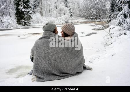 Giovane coppia coperta in coperta che si guarda l'un l'altro mentre seduto accanto al fiume ghiacciato nella foresta Foto Stock