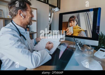 Medico di sesso maschile che parla del risultato della traccia del polso con il paziente su videochiamata tramite computer in ufficio Foto Stock