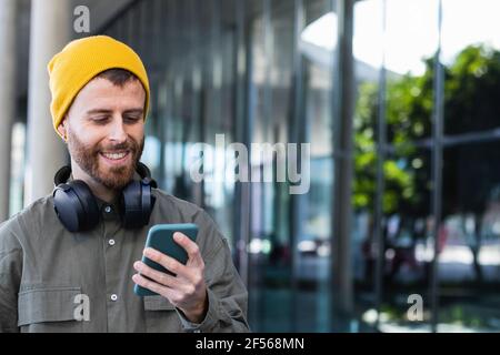 Sorridente bell'uomo con le cuffie che usano lo smartphone vicino all'edificio Foto Stock