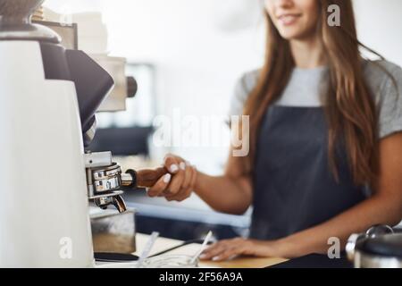 Primo piano di una barista femminile che macina il caffè per preparare una tazza di caffè perfetta in un bar luminoso in una giornata invernale soleggiata. Foto Stock