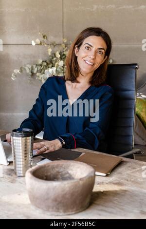 Donna matura sorridente seduta alla scrivania in ufficio Foto Stock