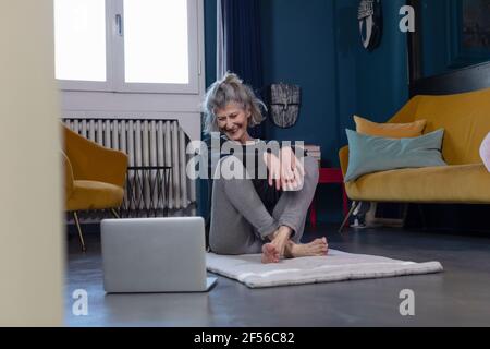 Donna anziana sorridente che guarda il tutorial sul computer portatile mentre si siede a. casa Foto Stock