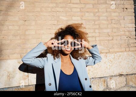 Sorridente giovane donna in occhiali da sole che gesturano mentre si trova contro il muro Foto Stock