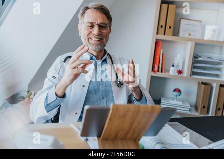 Medico maschile gesturing mentre si parla di videochiamata su digitale tablet presso l'ufficio del medico Foto Stock