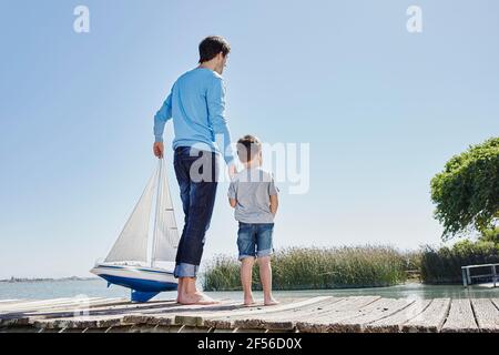 Uomo maturo con la barca a vela giocattolo che tiene la mano di ragazzo mentre in piedi contro il cielo Foto Stock