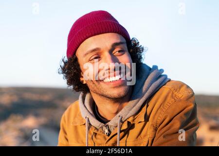 Uomo sorridente che indossa un cappello a maglia che guarda lontano mentre si siede all'aperto Foto Stock