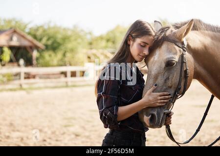 Cura, tenerezza e concetto di animali. Bella e spensierata tenera giovane donna in camicia controllata, appoggiata sul viso del cavallo, occhi stretti e sorridente dolcemente Foto Stock