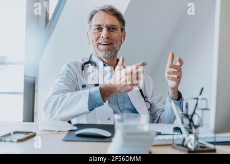 Sorridente medico maschile gesturing mentre si siede in ufficio medico Foto Stock