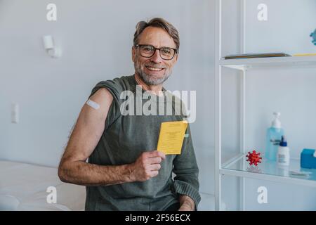 Uomo sorridente con bendaggio sul braccio che mostra il certificato di vaccinazione mentre seduta nella sala esame Foto Stock