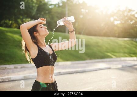 Giovane idolo di idoneità femminile che versa l'acqua sopra il suo corpo perfetto dopo un workout lungo e duro. Concetto di sport urbano. Foto Stock