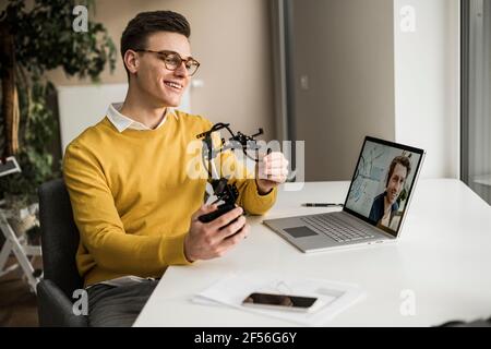 Sorridente ingegnere maschile con braccio robotico che partecipa alla videoconferenza mentre seduto alla scrivania Foto Stock