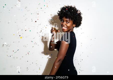 Donna con capelli ricci che mostra un flauto di champagne mentre si sta in piedi contro il bianco sfondo Foto Stock