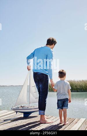 Padre con la barca a vela giocattolo che tiene la mano del figlio mentre si è in piedi sul molo Foto Stock