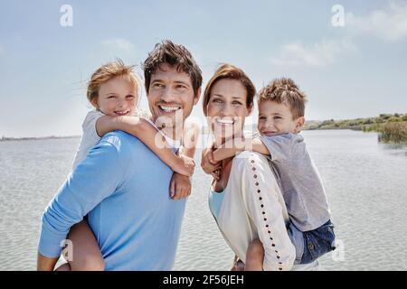 Sorridi genitori bambini piggybacking da lago in giornata di sole Foto Stock