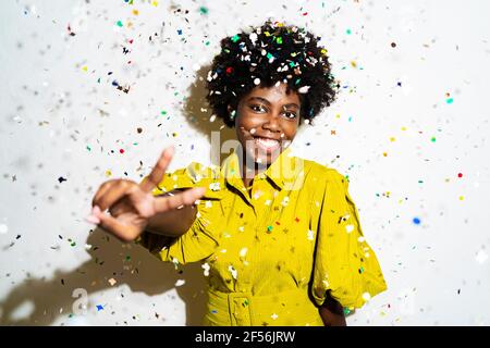 Donna allegra capelli ricci che mostra un segno di pace mentre si trova in mezzo confetti su sfondo bianco Foto Stock