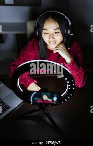 Sorridente influencer femmina che indossa le cuffie davanti all'anello illuminato leggero Foto Stock